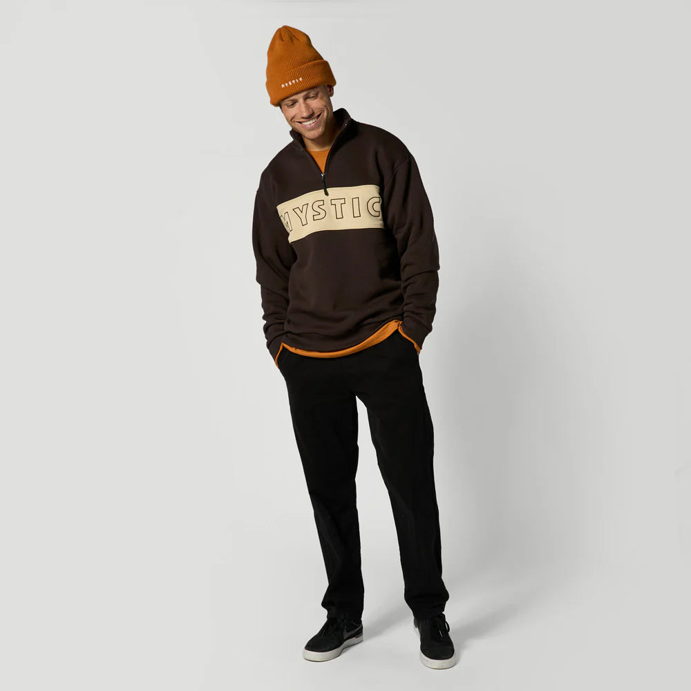 Person wearing a brown sweatshirt with 'Mystic' logo and orange beanie on a white background